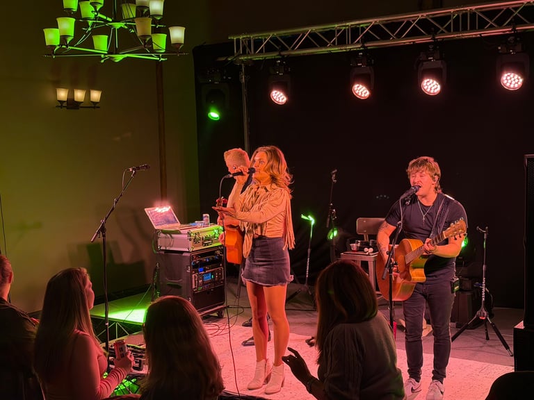 Band performing on stage with orange and pink stage lighting, featuring vocalist, keyboardist, and guitarist with audience members in foreground